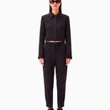 A model stands against a pale pink studio background wearing a black cropped long-sleeve collared jacket with a matching belt and black trousers, styled with black sneakers and sunglasses.