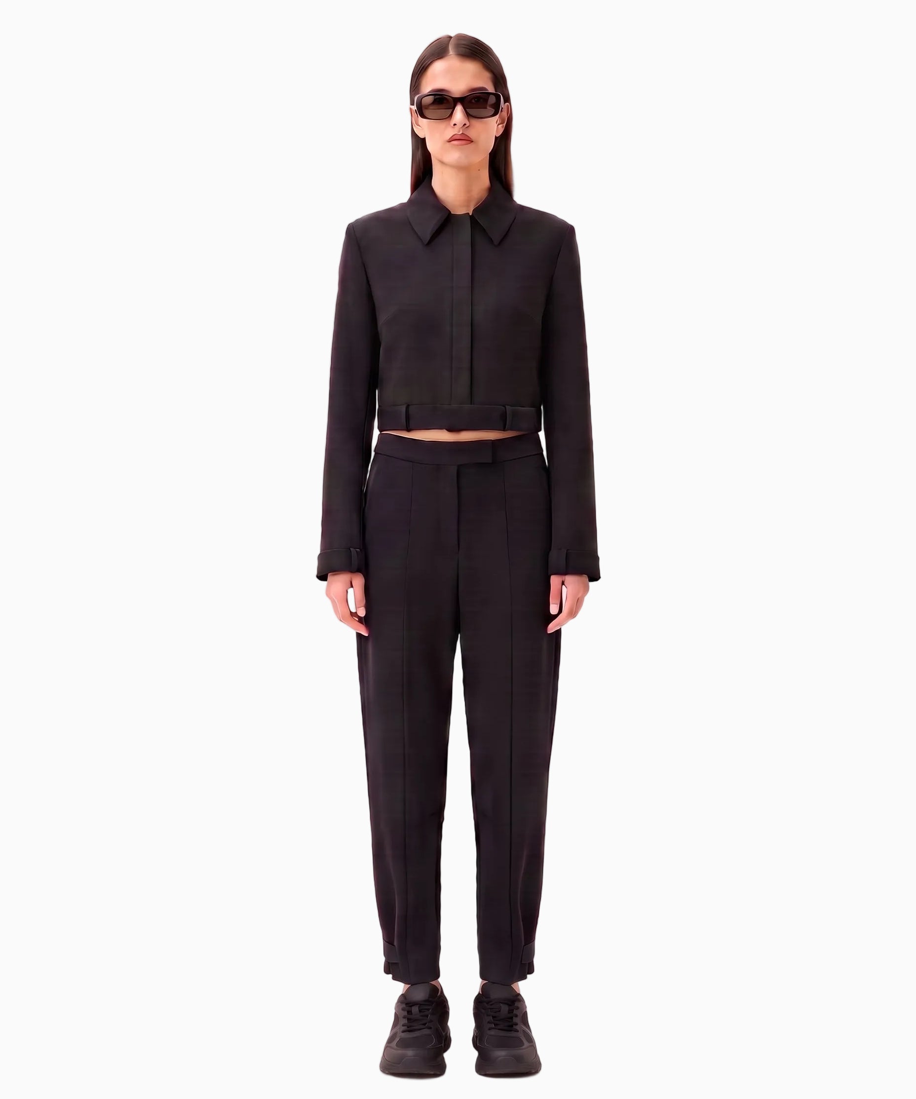 A model stands against a pale pink studio background wearing a black cropped long-sleeve collared jacket with a matching belt and black trousers, styled with black sneakers and sunglasses.