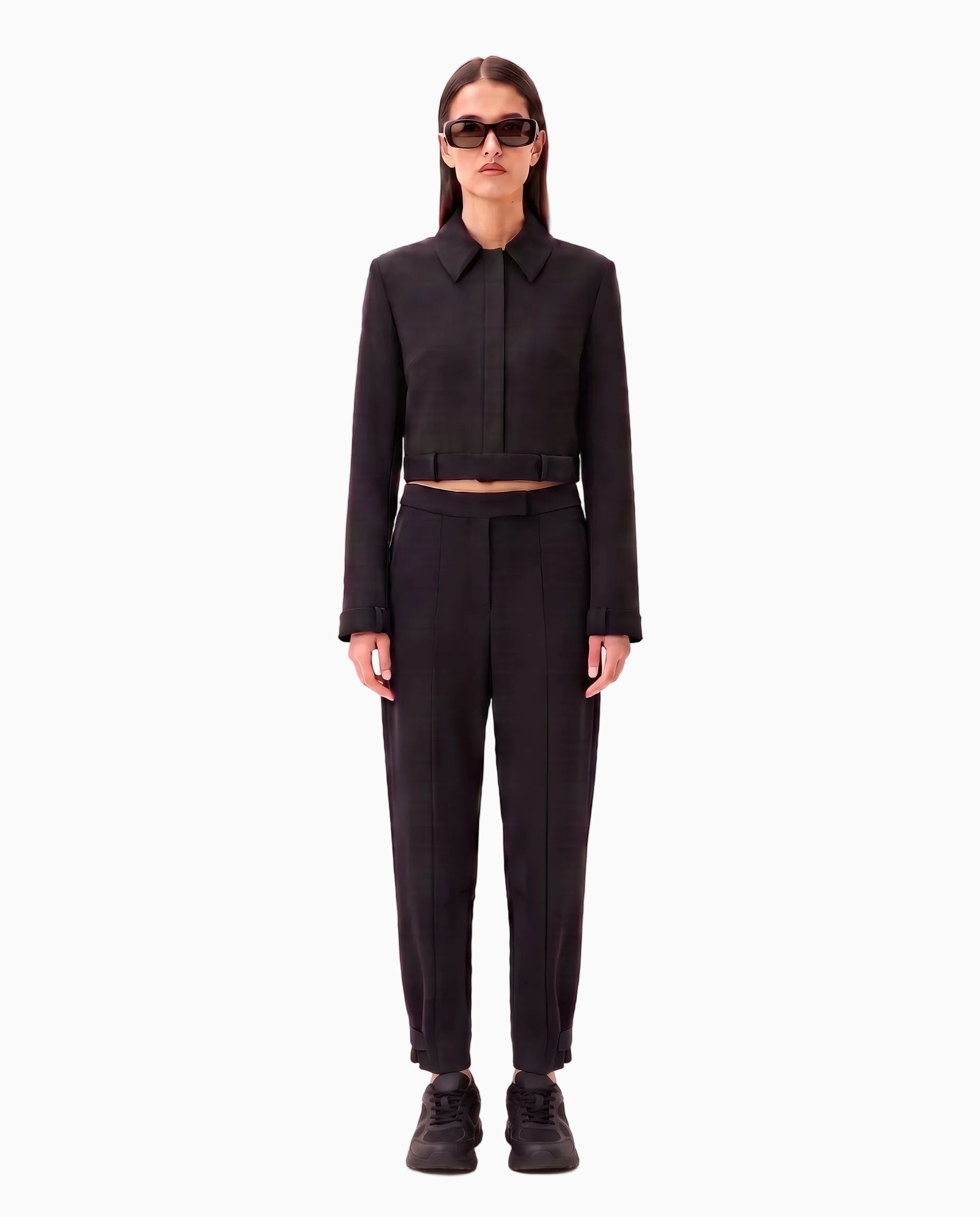 A model stands against a pale pink studio background wearing a black cropped long-sleeve collared jacket with a matching belt and black trousers, styled with black sneakers and sunglasses.